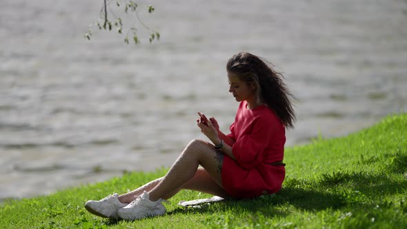 Young Lady with Smartphone on Shore of River in City at Summer Day Internet Addiction