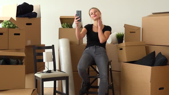 A Moving Woman Sits on a Chair in an Empty Apartment and Takes Selfies, Surrounded By Boxes