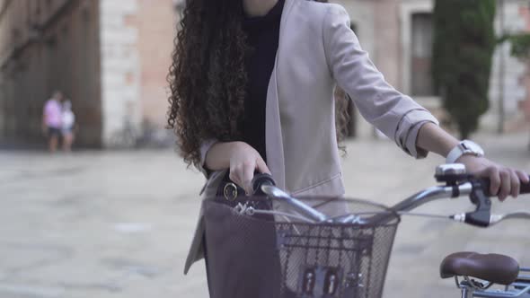 Pretty Woman With Long And Curly Hair Is Walking Along The Street With Bicycle