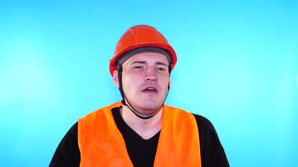 Male Construction Worker in Overalls Removing Medical Mask From Face on Blue Background