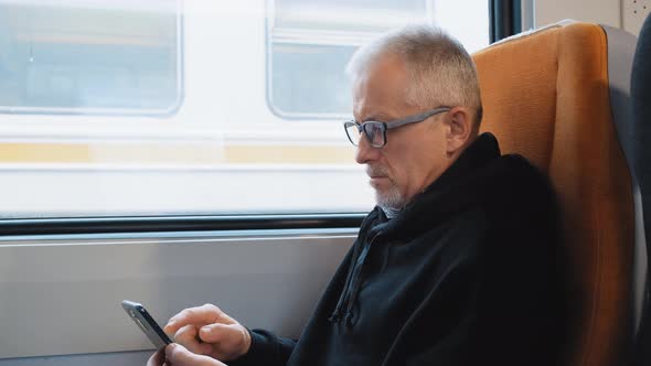 An Elderly Man is Sitting on a Train and Studying His Phone While Waiting for a Trip