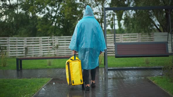 Back View Wide Shot Courier in Rain Coat Walking with Yellow Rucksack in Urban City Park Turning