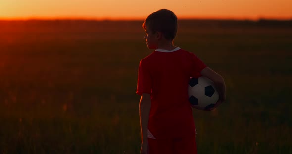 The Camera Follows the Boy Walking at Sunset in the Field with the Ball