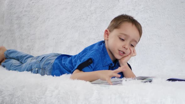 A Happy Child Happily Reads a Book Lying on a White Sofa. The Concept of Child Development