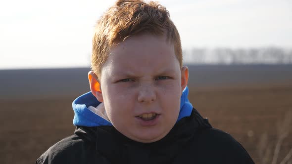 Young Redhaired Boy Looks Into Camera with Angry Facial Expression Against the Blurred Background of