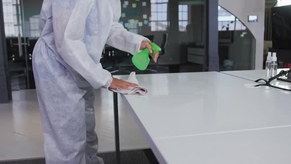 Midsection of cleaner wearing protective overalls disinfecting office and furniture