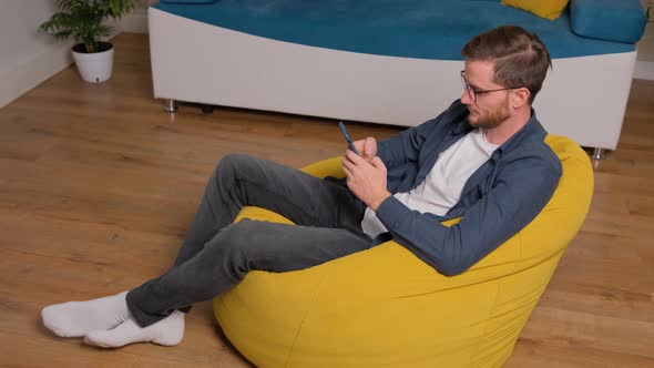 Young Man Sits on Bean Bag and Playing Games on Phone, at Home.