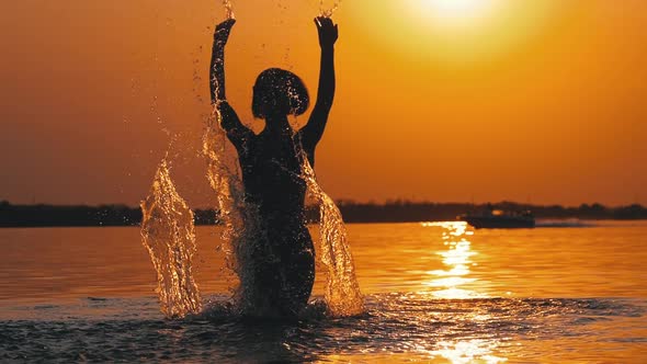 Silhouette of Woman at Sunset Raises Hands Up and Creating Splashes of Water. Slow Motion