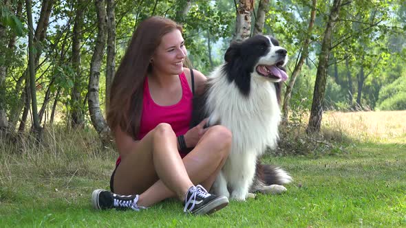 A Young Beautiful Woman Sits on Grass in a Meadow with a Border Collie and Smiles at the Camera