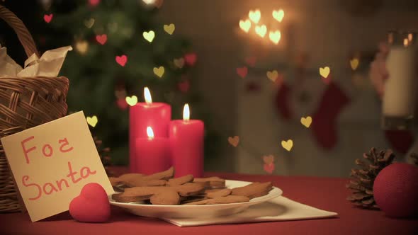 Girl Putting Glass of Milk for Santa Near Burning Candles and Honey Cookies