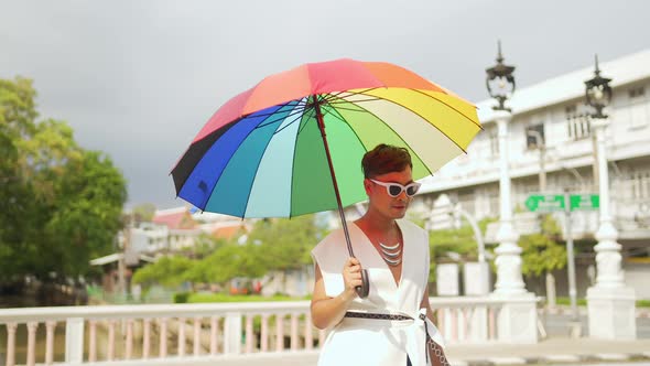 Asian lgbtq guy wearing woman clothes with sunglasses holding rainbow umbrella walking down street.