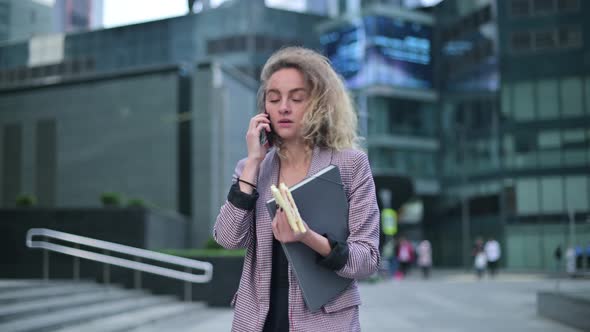 woman strides rapidly, talking on the phone and holding a sandwich in her hand