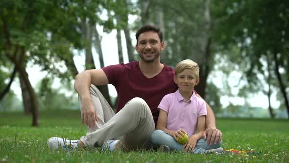 Boy Sitting With Father in Park, Showing Fresh Apple Into Camera, Healthy Food