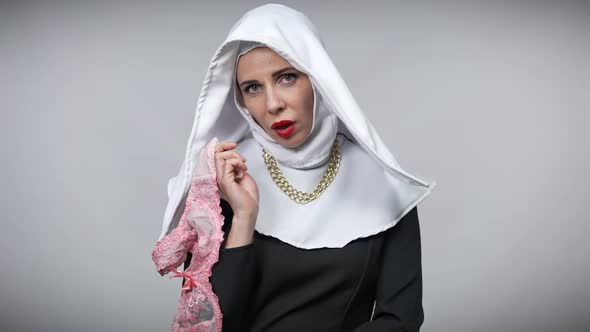 Seductive Woman in Nun Costume Holding Pink Lace Bra Looking at Camera Posing at Grey Background
