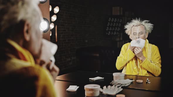 Clown Removes Makeup with Napkin Sitting Against Mirror