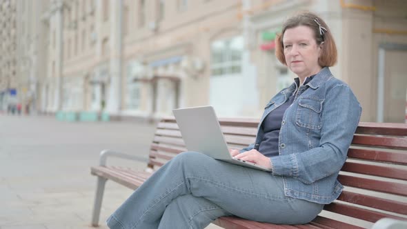 Old Woman with Laptop Smiling at Camera While Sitting Outdoor on Bench