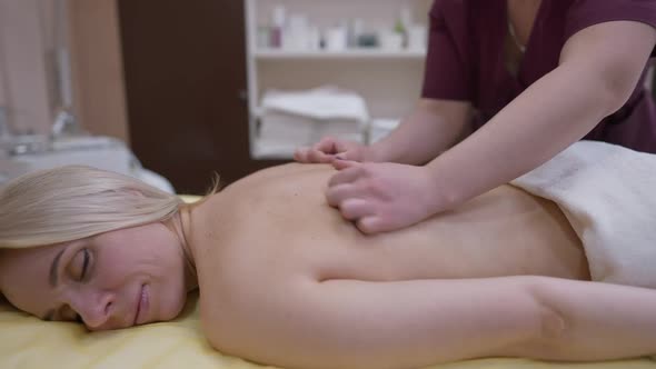 Side View Smiling Woman with Closed Eyes Lying in Spa Salon As Massagist Rubbing Back in Slow Motion