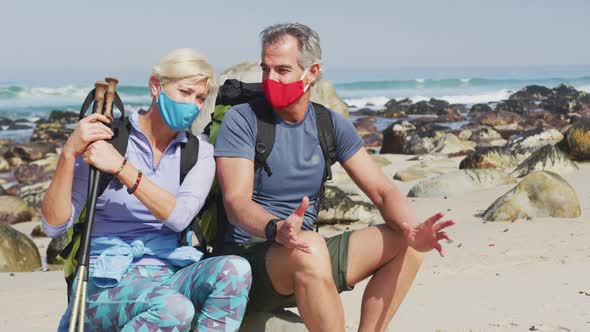 Senior hiker couple wearing face masks with backpacks and hiking poles talking sitting on rock