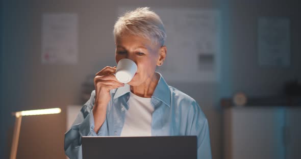 Mature Businesswoman Working on Laptop Holding Coffee Cup at Night in Office