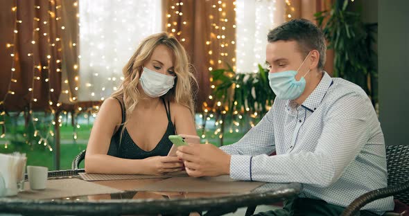 Couple with Surgical Masks Uses Phones Sitting at Table