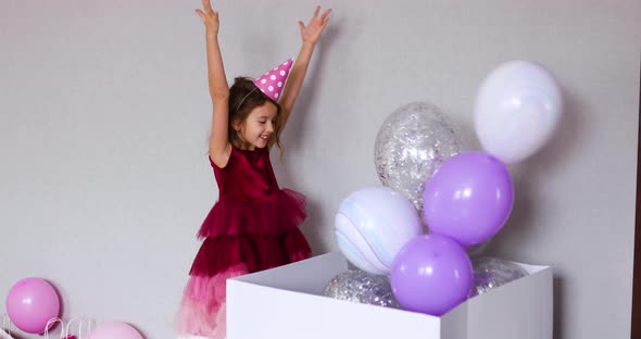 Joyful little girl in pink dress and hat open big present gift box with balloons 