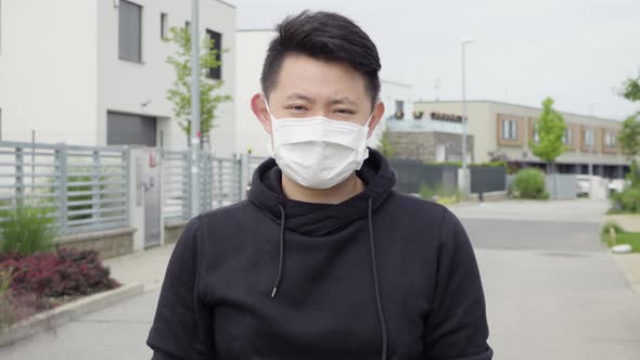 A Young Asian Man in a Face Mask Looks at the Camera in an Empty Suburban Area