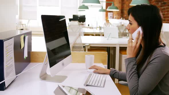 Female executive talking on mobile phone while working on computer at desk