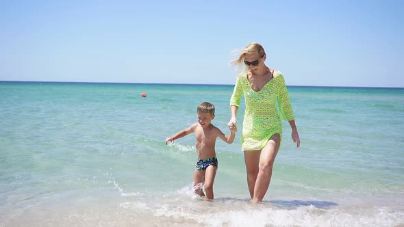 Little Boy Holds His Mother's Hand and Creates Sea Spray on a Sandy Beach on a Sunny Day