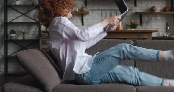 Side View of Excited Redhaired Young Woman Sits at on Sofa at Laptop Computer Working in Living Room