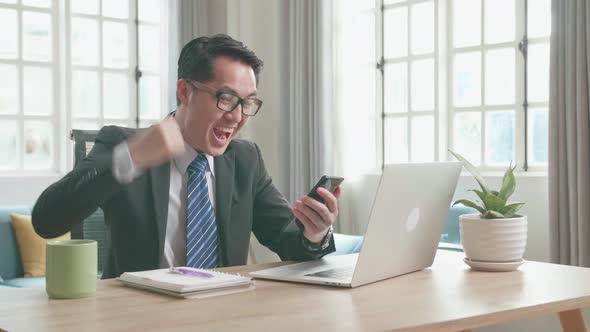 Happy Asian Businessman Looking At Mobile Phone While Using Computer For Working At Home