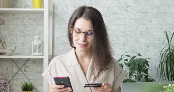 Smiling Young Woman with Glasses Holding Credit Card and Smartphone Sitting on at Home. Happy Female