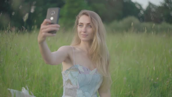 Middle Shot of Beautiful Woman in Elegant Dress Sitting on Green Meadow and Taking Selfie. Portrait