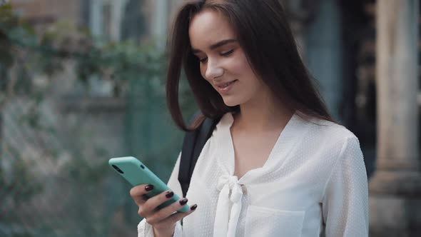 Close Up Shot of Beautiful Student Girl with Bag on Her Shoulder Using Her Modern Smartphone. Young