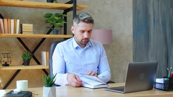 Focused man at home office in wireless earphones writes information notebook looks laptop computer