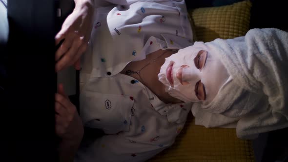 Young Woman in Cosmetic Fabric Mask and White Towel on Her Head, Is Working on Laptop, Watching on