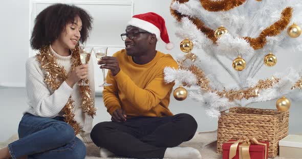 Young Lovely Afro American Couple Sitting and Dancing Moving Near Christmas Tree and Drinking Glass