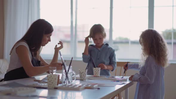 Group of Kids and Their Teacher Leaning Over Table with Watercolor Paints and Painting Selfmade Clay