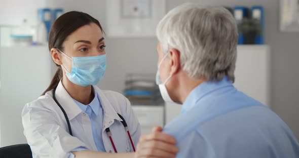 Close Up Female Doctor Wearing Mask Touching Senior Patient Shoulder in Clinic Office