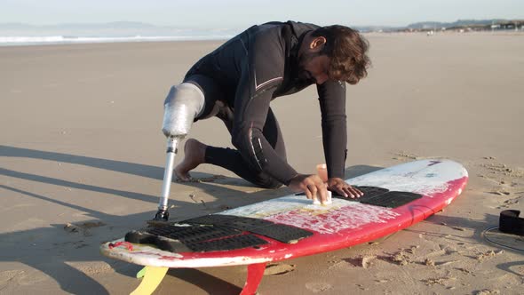 Long Shot of Surfer with Disability Waxing Surfboard on Beach