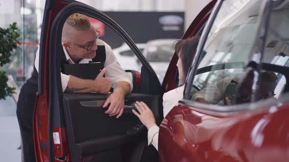 Smiling Professional Male Car Dealer with Tattoo and Mohawk Haircut Standing at Open Car with Female