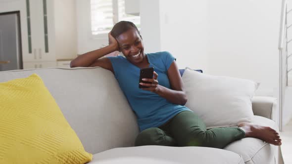 Happy african american mother sitting on sofa using smartphone and laughing