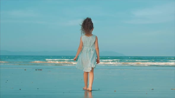 Curly Little Girl in White Dress Walking on the Beach at Sunset