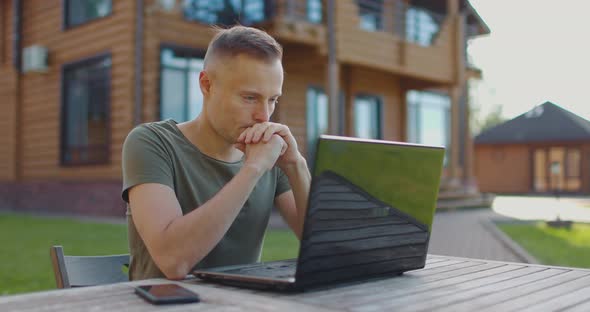 Young Serious Man Watching Webinar on Laptop While Sitting at Table in Courtyard of House