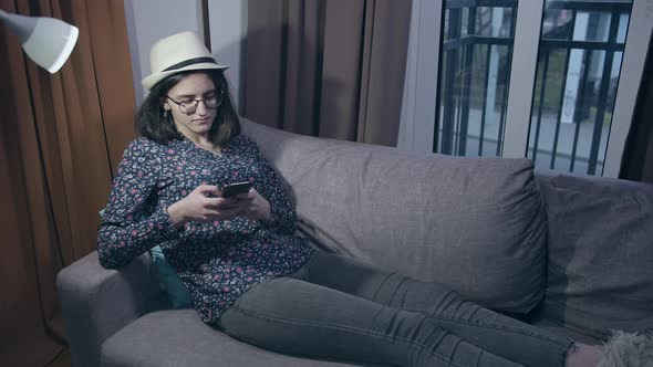 A Girl Student in Glasses and a Hat Reads Messages in Her Mobile Phone While Sitting on the Couch