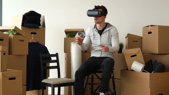 A Moving Man Sits on a Chair in an Empty Apartment and Uses VR Headset, Surrounded By Boxes