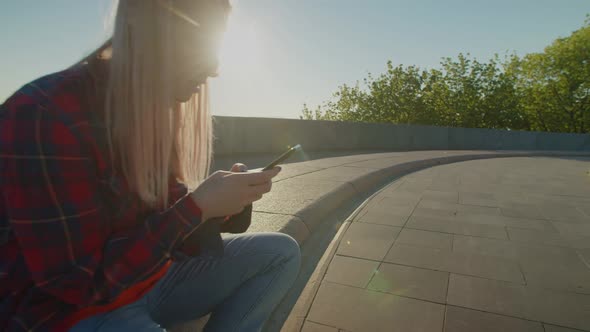 Cute Skateboarder Sitting on Skateboard Using Mobile Phone at Daybreak