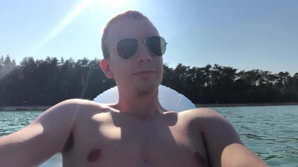 Young Man Relax on Inflatable Deck Chair on Lake During Sunny Day and Smiles To Camera