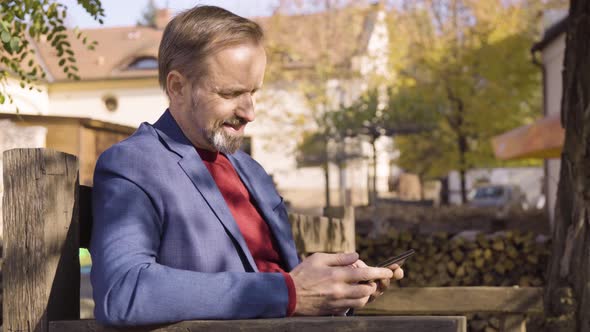 A Middleaged Caucasian Man Works on a Smartphone with a Smile As He Sits on a Bench