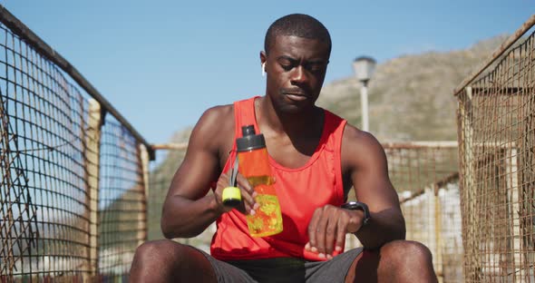 African american man sitting, drinking from water bottle, taking break in exercise outdoors