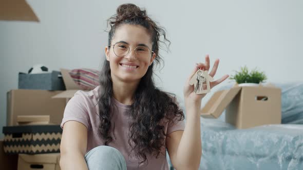 Joyful Mixed Race Lady Raising Hand with House Key Indoors in Room Full of Cardboard Boxes